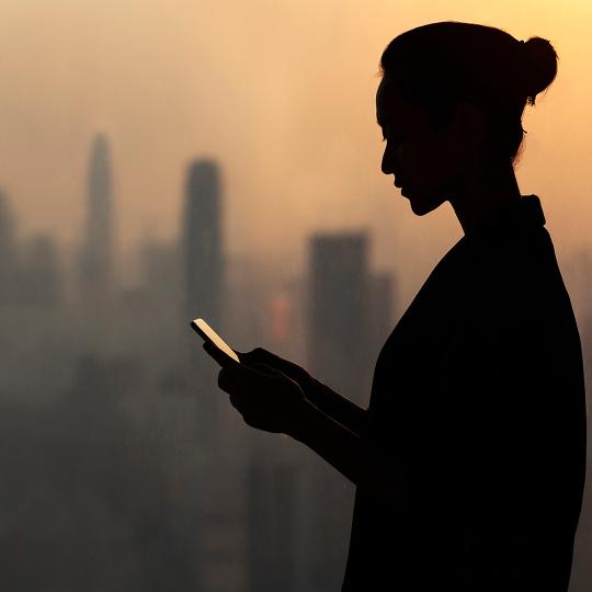 Silhouette of young woman using smartphone next to window with cityscape in background.