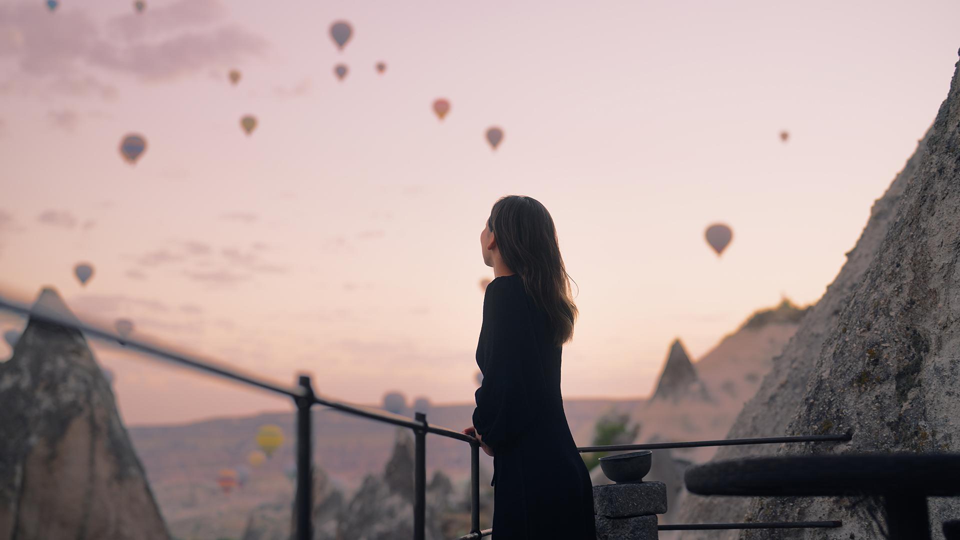 A female tourist is enjoying watching hot air balloons flying in the sky at the rooftop of the hotel where she is staying during her vacation.